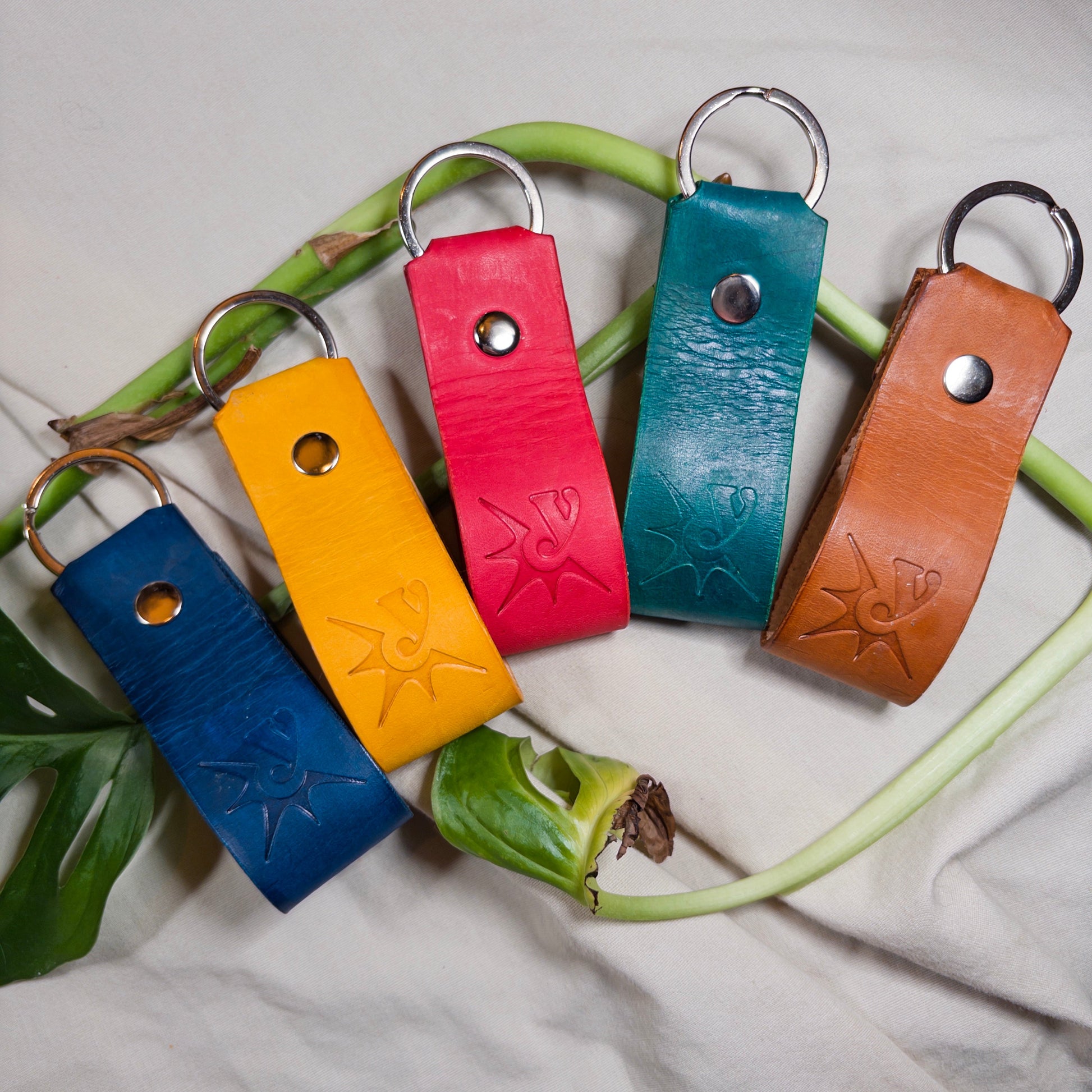 Five colorful leather keychains on a white fabric background with green leaves.