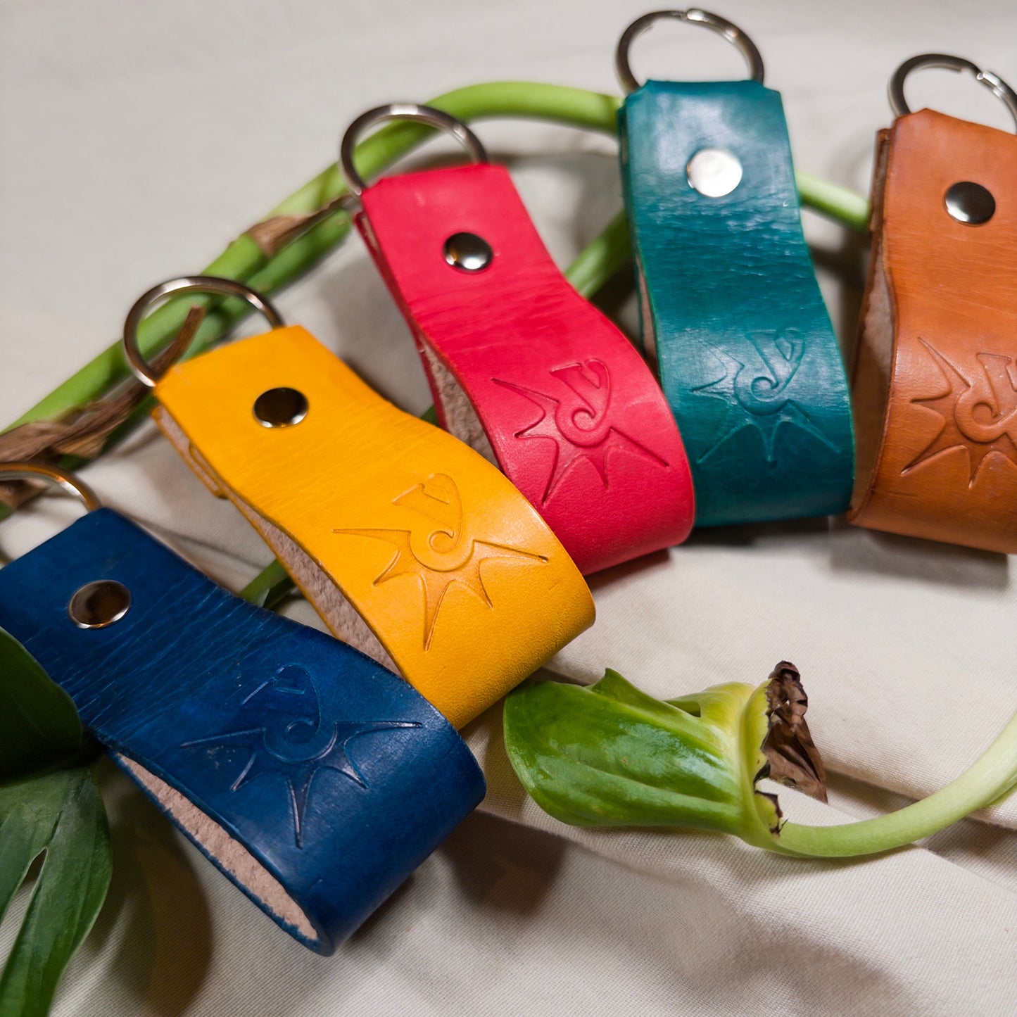 Five colorful leather keychains on a light fabric background with green leaves.